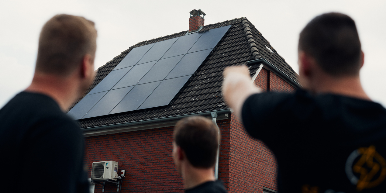 drei Personen blicken auf ein Haus mit Photovoltaikanlage auf dem Dach
