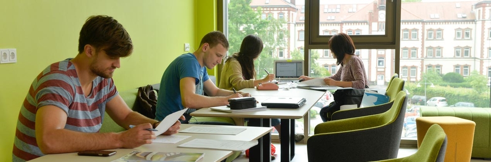 Vor einem Fenster mit Blick auf das alte Hauptgebäude des Campus sitzen mehrere Studierende an Tischen und lernen.