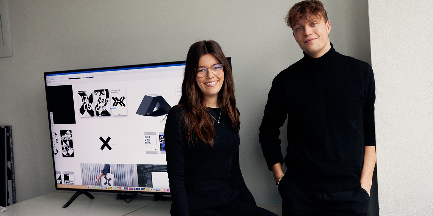 Pauline und Paul stehen nebeneinander vor einem großen Bildschirm auf dem man Designentwürfe von der CXI sieht